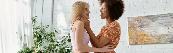 A multicultural lesbian couple stands in their modern apartment, smiling at each other. — Stock Photo