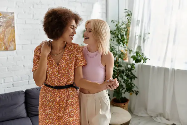 Two women in an apartment - one with an curly hair and floral dress, the other with blonde hair and pink top, hold hands and smile at each other — Stock Photo