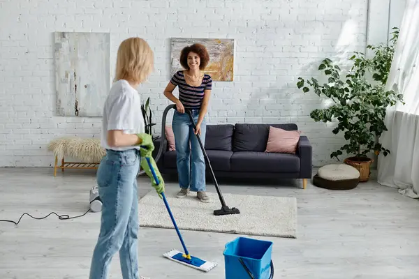 Duas mulheres, uma esfregando e a outra aspirando, limpam seu apartamento moderno juntas.. — Fotografia de Stock