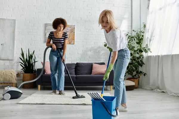 Um casal lésbico multicultural limpa seu apartamento moderno juntos, com uma mulher aspirando o tapete e a outra limpando o chão. — Fotografia de Stock