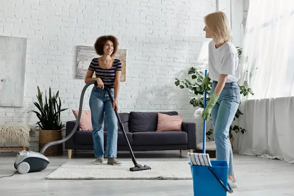 Duas mulheres de raças diferentes limpam o seu apartamento em conjunto - um aspirador em calça, o outro esfregona em uma camiseta branca — Fotografia de Stock