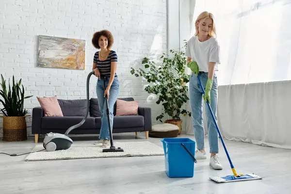 Um casal lésbico multicultural limpa seu apartamento moderno juntos, um aspirando e o outro esfregando. — Fotografia de Stock