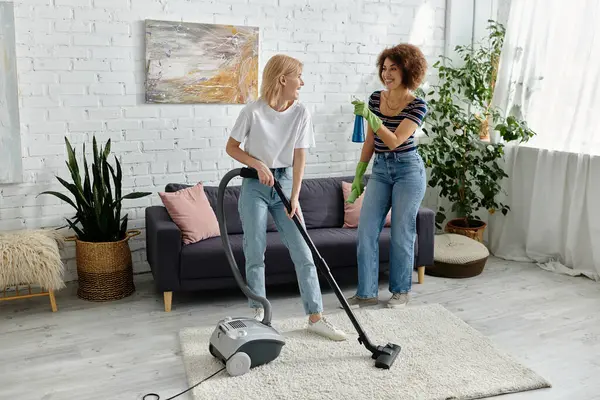 Duas mulheres, uma aspirando e a outra pulverizando limpador, rir como eles limpam seu apartamento moderno. — Fotografia de Stock