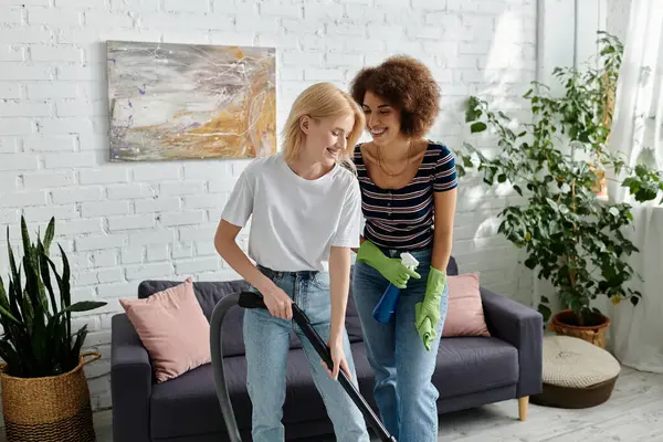 Duas mulheres limpam o apartamento moderno juntas, sorrindo e rindo. Um vácuo enquanto o outro usa spray de limpeza e pano — Fotografia de Stock