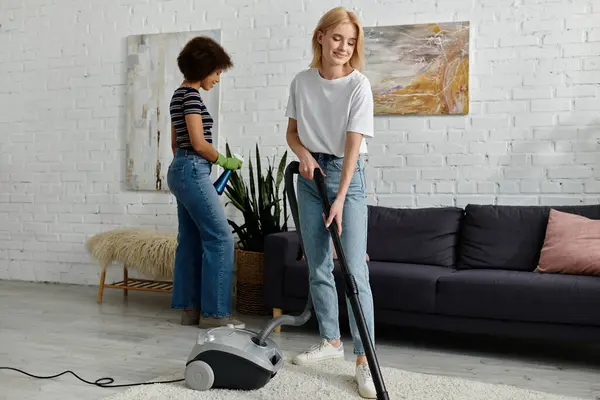 Um casal de lésbicas limpa seu apartamento moderno juntos, com uma mulher aspirando o tapete enquanto a outra segura uma garrafa de spray. — Fotografia de Stock