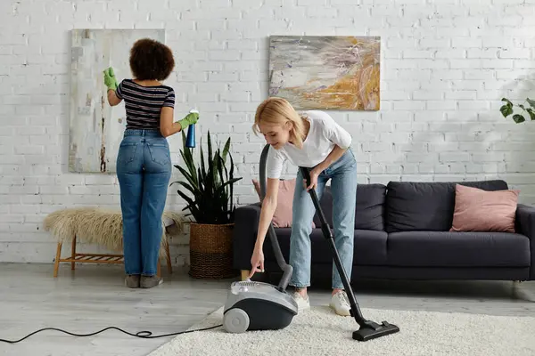 Um casal lésbico multicultural limpa seu apartamento moderno, um espanando obras de arte, enquanto o outro aspira o tapete. — Fotografia de Stock
