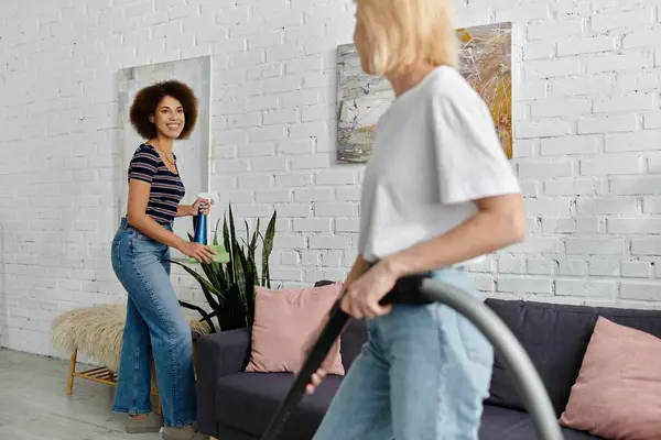 Um casal de lésbicas limpa seu apartamento juntos, um segurando um spray de limpeza e o outro com um aspirador de pó.. — Fotografia de Stock