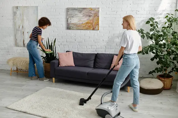 Um casal lésbico multicultural limpa seu apartamento moderno juntos, um espanando e o outro aspirando. — Fotografia de Stock