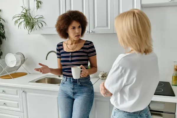 Un couple de lesbiennes a une discussion tendue dans leur cuisine, une tasse de café oubliée dans la main de la femme à la chemise rayée. — Photo de stock