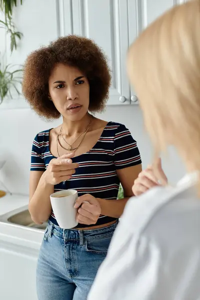 Une femme écoute attentivement son partenaire, une femme blonde, tenant une tasse de café dans une cuisine, les deux regardant mécontents — Photo de stock