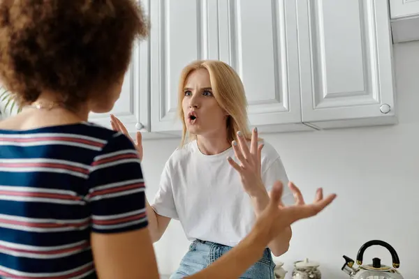 Deux femmes, l'une aux cheveux blonds et l'autre aux cheveux bruns bouclés, se disputent dans une cuisine. La femme aux cheveux blonds fait un geste avec les deux mains. — Photo de stock