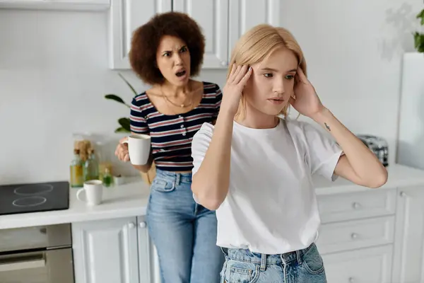 Un couple de lesbiennes se tient dans la cuisine, l'une tenant une tasse, l'autre tenant sa tête, comme ils se disputent. — Photo de stock