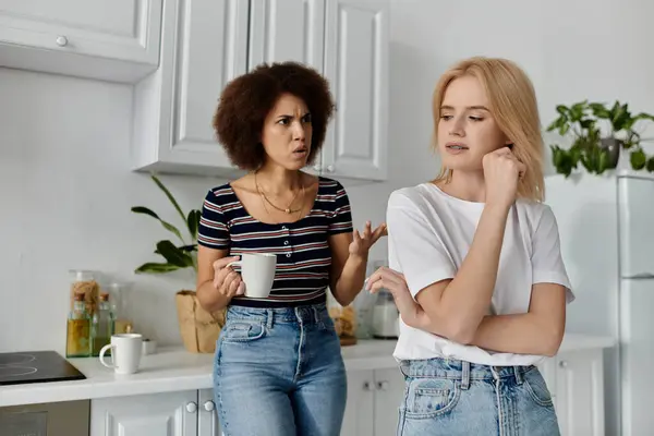 Un couple de lesbiennes se dispute dans leur cuisine, l'une tenant une tasse. La femme de gauche parle tandis que la femme de droite écoute — Photo de stock