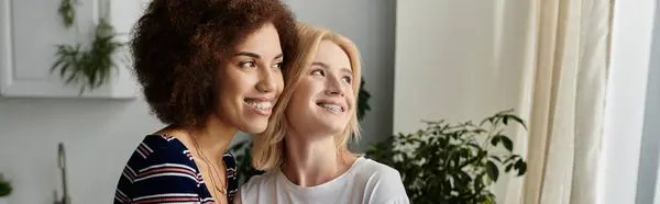 A young lesbian couple stands in the kitchen, facing a window. They are both looking away from the camera — Stock Photo