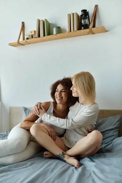 A multicultural lesbian couple cuddles on a bed, dressed in comfy homewear, sharing a tender moment together. — Stock Photo