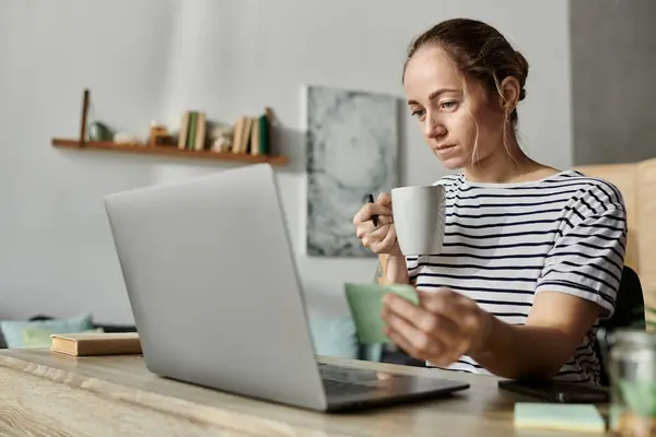 A woman with vitiligo sips coffee, focused on her laptop screen during a cozy morning. — Stock Photo