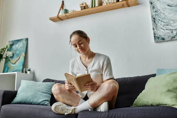 Uma jovem mulher gosta de um livro enquanto se senta pacificamente em um sofá aconchegante. — Fotografia de Stock