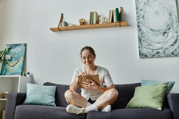 A cheerful woman holds a book while sitting cross-legged on a sofa. — Stock Photo