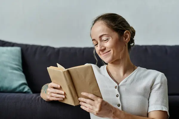Una mujer con vitiligo sonríe sumergida en un libro en un cómodo sofá. - foto de stock