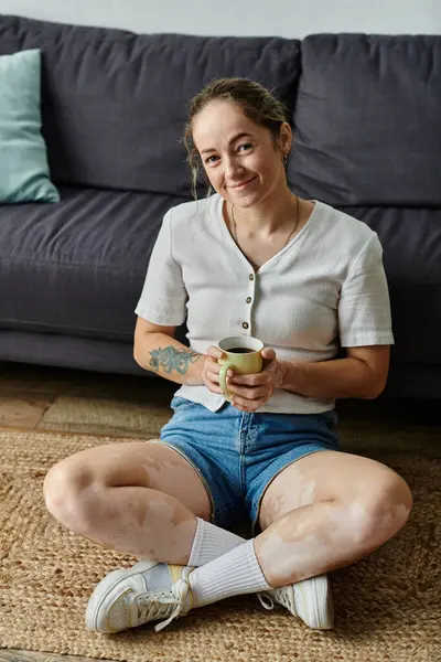 Una mujer alegre se sienta con las piernas cruzadas, bebiendo té y sonriendo calurosamente en un espacio acogedor. — Stock Photo