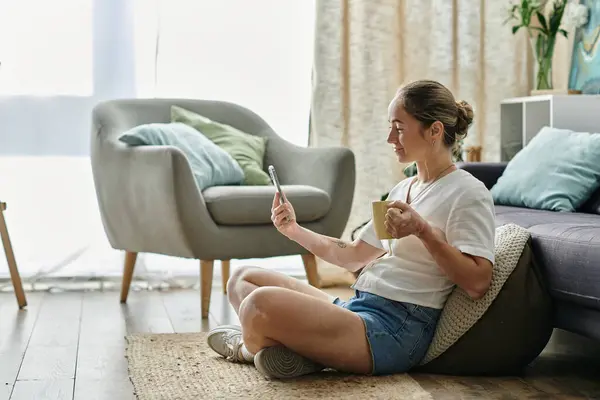 A young woman relaxes on the floor, sipping tea while engaging with her smartphone. — Stock Photo