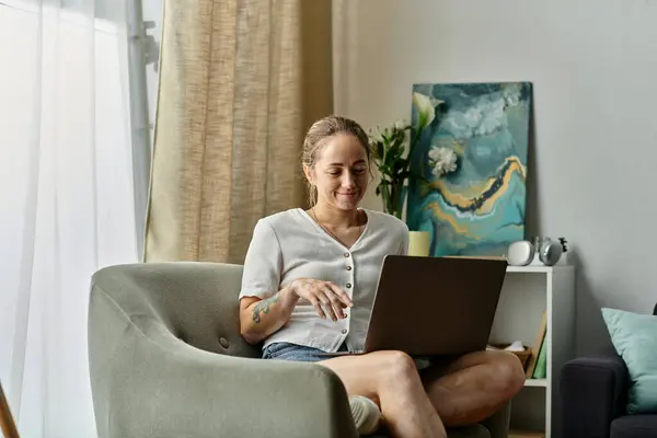 Mujer sonriente se involucra con su computadora portátil mientras se sienta cómodamente en el interior. - foto de stock