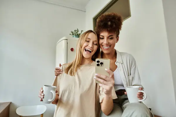 Two partners share laughter while looking at their phone, holding warm drinks. — Stock Photo