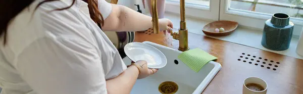 A beautiful plus size woman joyfully washes dishes in her cozy kitchen. — Stock Photo
