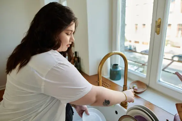 A beautiful plus size woman happily washes dishes by the sunny kitchen window. — Stock Photo