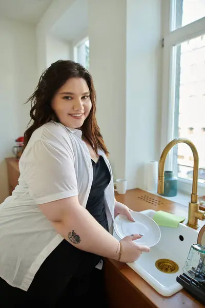 Eine Plus-Size-Frau lächelt beim Geschirrspülen in ihrer hellen Küche. — Stockfoto