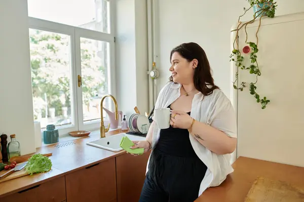 A cheerful woman enjoys her morning beverage while standing in a sunny kitchen. — Stock Photo