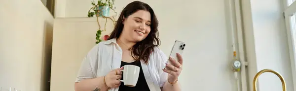 Une femme gaie plus la taille se détend tout en vérifiant son téléphone et en sirotant du café. — Photo de stock