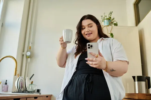 Smiling woman sips coffee and checks her phone in a cozy kitchen setting. — Stock Photo