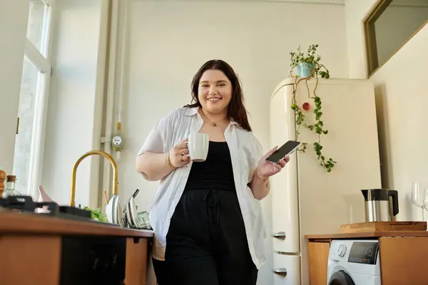 Relaxing at home, a plus size woman holds a mug and a phone while smiling. — Stock Photo