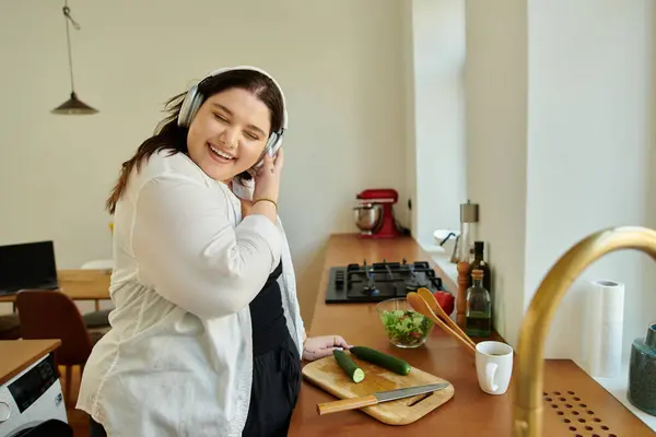 Una mujer alegre prepara una comida fresca, usando auriculares y sonriendo. - foto de stock