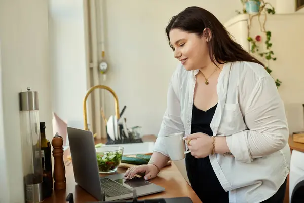 A cheerful woman enjoys her drink while working on her laptop at home. — Stock Photo