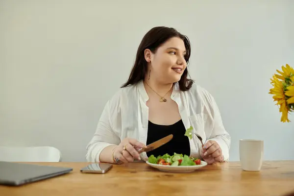 A plus size woman smiles while savoring a vibrant salad at her dining table. — Stock Photo