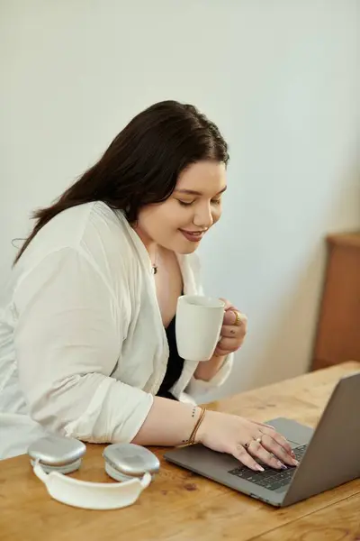 Uma mulher adorável gosta de seu café enquanto trabalha em um laptop em casa. — Fotografia de Stock