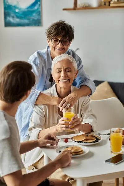 Loving lesbian parents enjoy breakfast with their adult son, sharing laughter and conversation at home. — Stock Photo