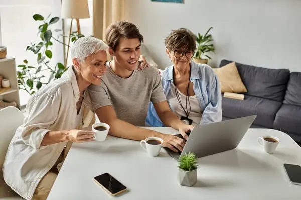 Lesbian parents and their adult son share laughter and warm moments over coffee while using a laptop. — Stock Photo