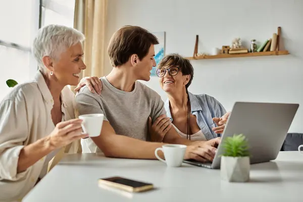 Lesbian parents share laughter and love with their adult son over coffee and a laptop in a sunny space. — Stock Photo