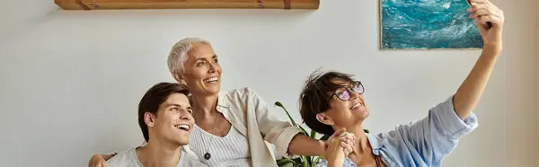 Lesbische Eltern und ihr erwachsener Sohn lachen, während sie ein Selfie in einem hellen, einladenden Wohnraum machen. — Stockfoto