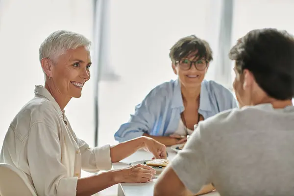 Lesbian parents share a joyful breakfast moment with their adult son at a sunlit kitchen table. — Stock Photo