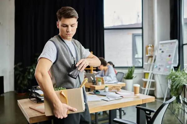 Jeune homme dévoué emballant ses articles pendant la mise à pied, collègues sur toile de fond. — Photo de stock