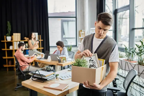 Junger gut aussehender Mann packt während der Entlassung seine Sachen, Kollegen im Hintergrund. — Stockfoto