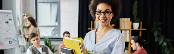 Merry african american woman packing her items during lay off, colleagues on backdrop. — Stock Photo