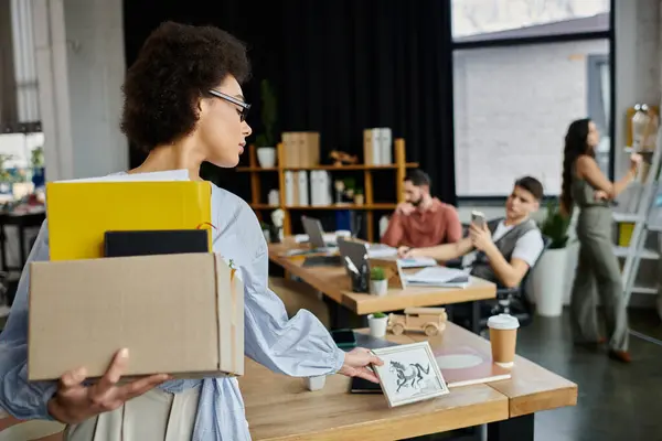 Stilvolle afrikanisch-amerikanische Frau packt während der Entlassung ihre Sachen, Kollegen im Hintergrund. — Stockfoto