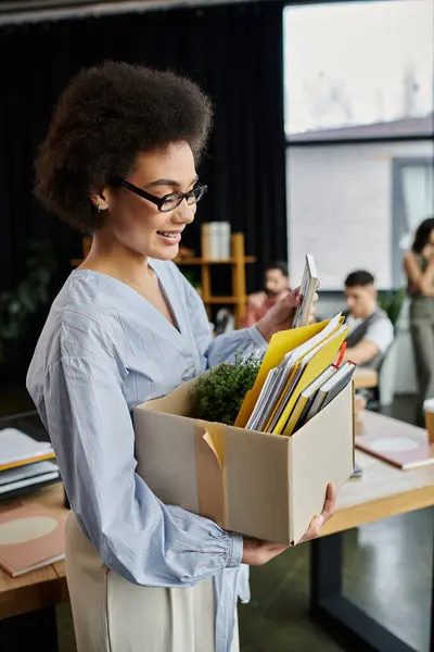 African american woman in chic attire packing her items during lay off, colleagues on backdrop. — Stock Photo