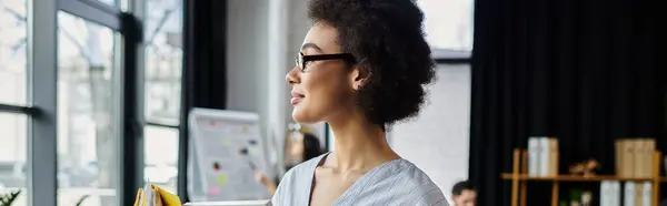 Professionale donna afroamericana imballare i suoi oggetti durante licenziare, colleghi sullo sfondo. — Foto stock