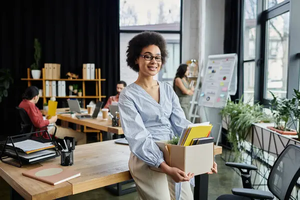 Positiva donna afroamericana imballare i suoi oggetti durante il licenziamento, colleghi sullo sfondo. — Stock Photo